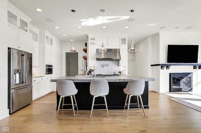 kitchen featuring an island with sink, light wood-style flooring, stainless steel appliances, a brick fireplace, and exhaust hood