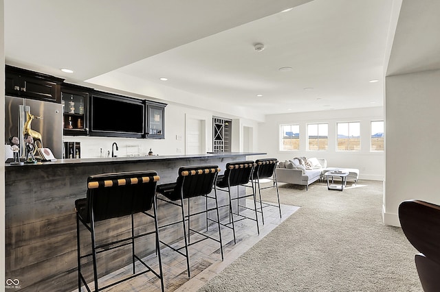 kitchen with light colored carpet, a breakfast bar, dark cabinetry, freestanding refrigerator, and glass insert cabinets