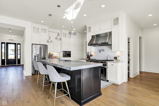 kitchen featuring light wood-style flooring, a kitchen island with sink, a kitchen breakfast bar, appliances with stainless steel finishes, and wall chimney exhaust hood