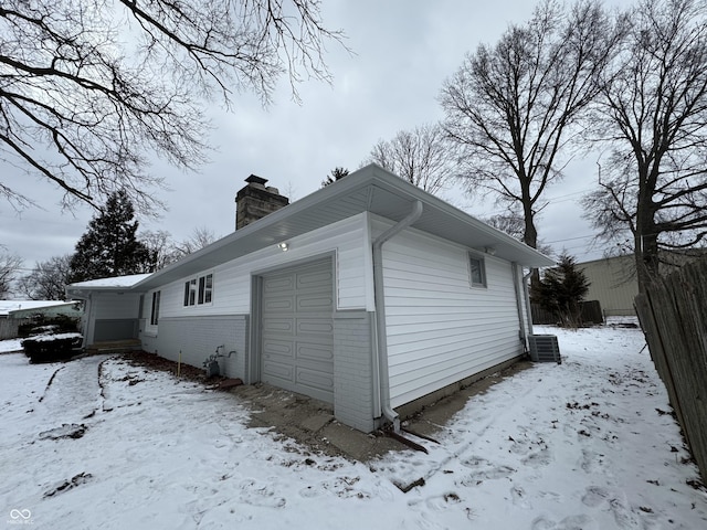 snow covered property with a garage, brick siding, a chimney, and central AC unit