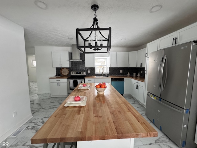 kitchen featuring marble finish floor, decorative light fixtures, stainless steel appliances, white cabinetry, and wall chimney range hood