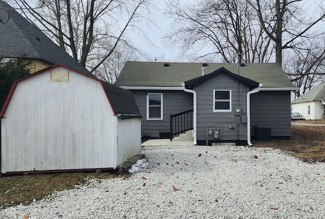 snow covered property with a storage shed, a shingled roof, cooling unit, and an outdoor structure