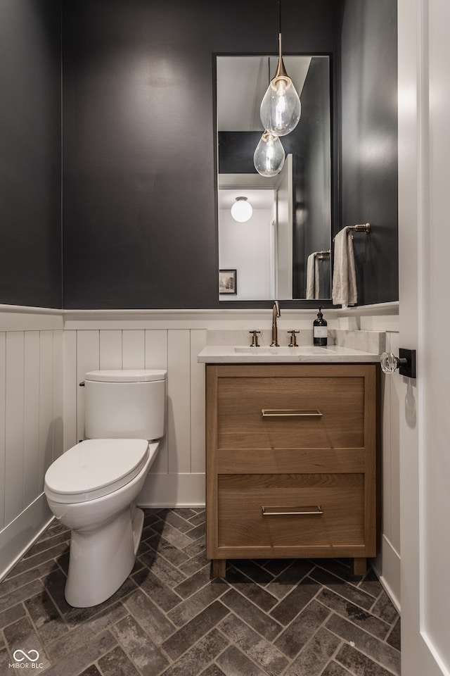 bathroom featuring toilet, brick floor, a wainscoted wall, and vanity