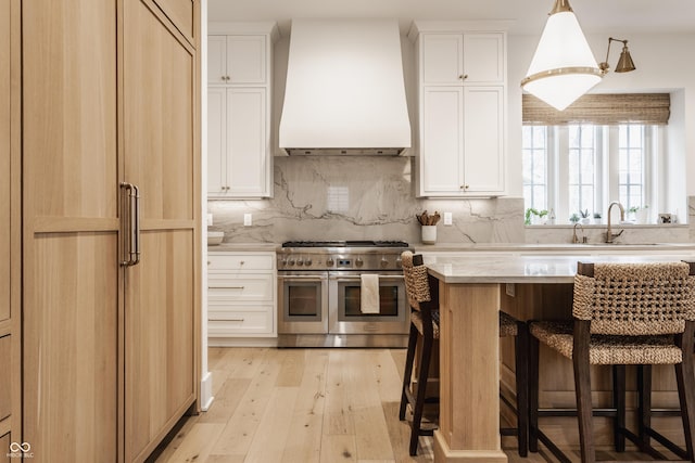 kitchen featuring range with two ovens, premium range hood, backsplash, light wood finished floors, and a kitchen bar