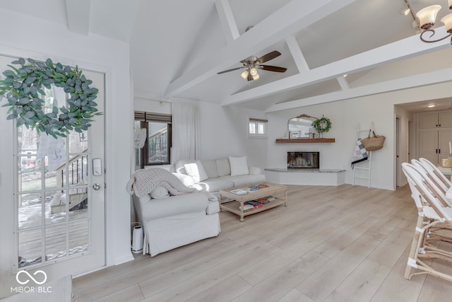 living area with vaulted ceiling with beams, a glass covered fireplace, light wood-style flooring, and a ceiling fan