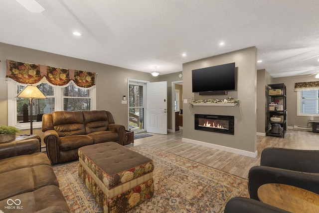 living room featuring a textured ceiling, recessed lighting, baseboards, light wood-type flooring, and a glass covered fireplace