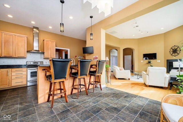 kitchen with arched walkways, backsplash, stainless steel electric stove, wall chimney exhaust hood, and dark countertops