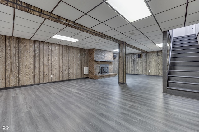 finished basement featuring a paneled ceiling, stairway, a brick fireplace, wood walls, and wood finished floors