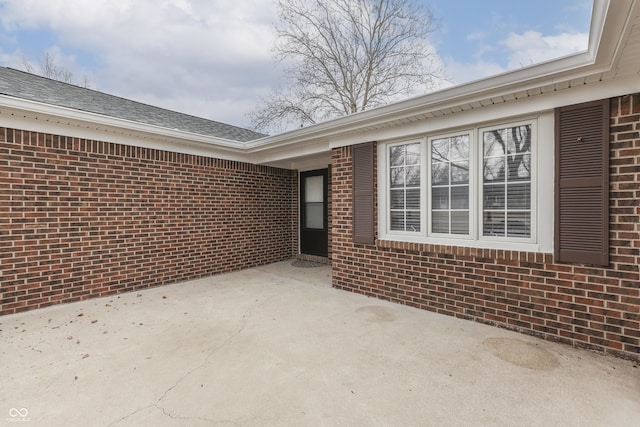 view of exterior entry with a shingled roof, brick siding, a patio, and driveway