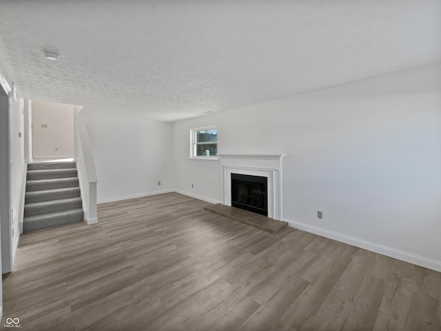 unfurnished living room featuring a fireplace with raised hearth, a textured ceiling, wood finished floors, baseboards, and stairway