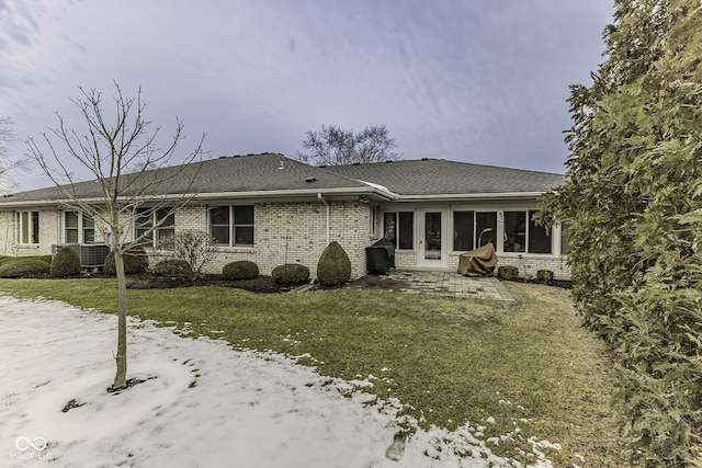 snow covered back of property featuring a patio, cooling unit, brick siding, a shingled roof, and a lawn