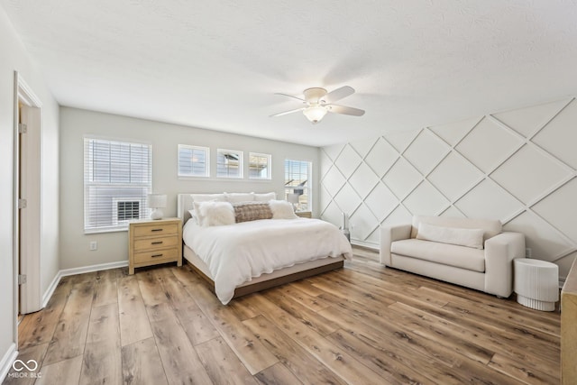 bedroom with a textured ceiling, light wood-style flooring, an accent wall, a ceiling fan, and baseboards