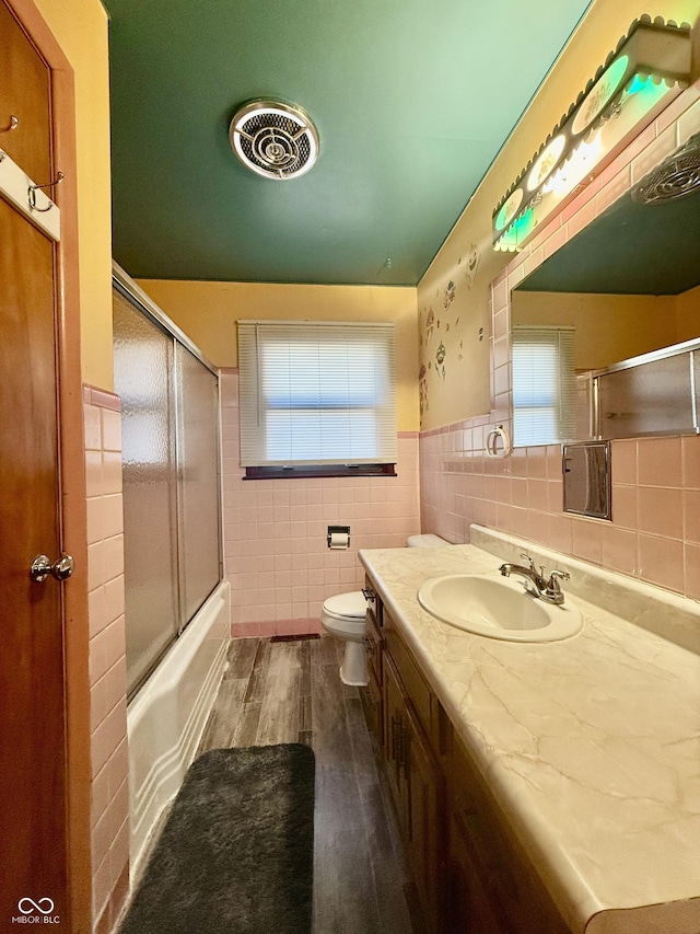 bathroom featuring toilet, bath / shower combo with glass door, wood finished floors, visible vents, and tile walls