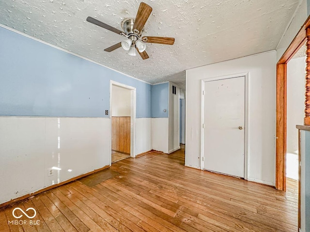 empty room featuring a wainscoted wall, light wood-style floors, ornamental molding, a ceiling fan, and a textured ceiling