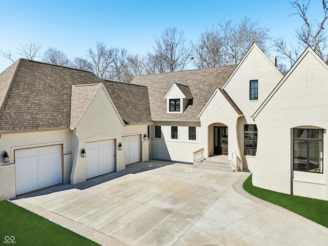 french country home with a garage, concrete driveway, roof with shingles, and brick siding