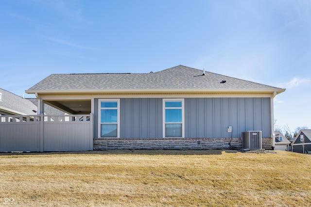 exterior space with board and batten siding, cooling unit, roof with shingles, and a lawn