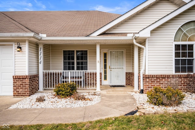 doorway to property with an attached garage, covered porch, roof with shingles, and brick siding