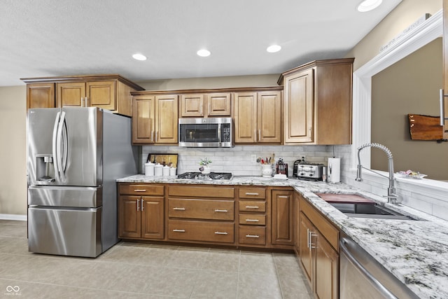 kitchen featuring tasteful backsplash, appliances with stainless steel finishes, brown cabinetry, a sink, and light stone countertops