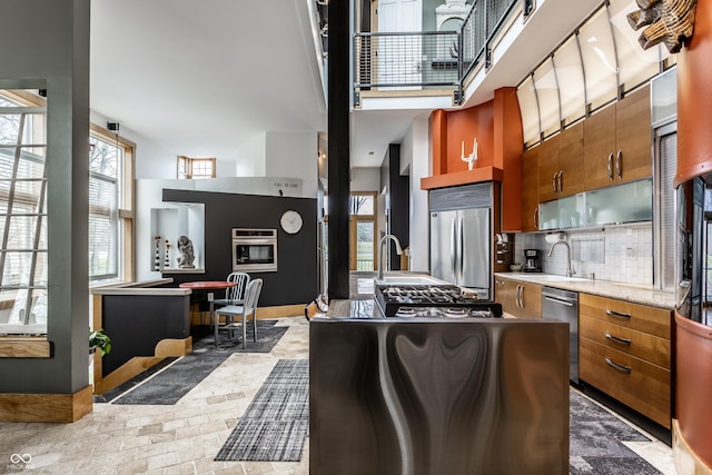 kitchen with brown cabinetry, a sink, decorative backsplash, stainless steel appliances, and a towering ceiling