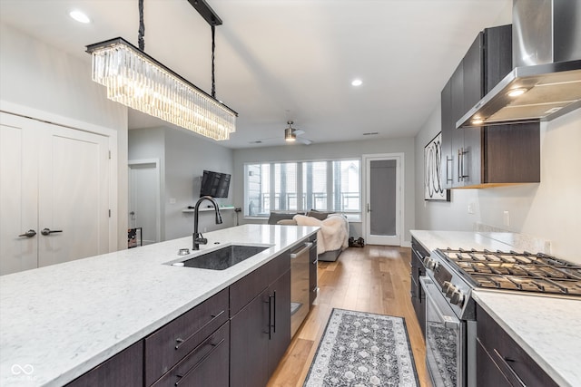 kitchen featuring dark brown cabinetry, wall chimney exhaust hood, appliances with stainless steel finishes, light wood-style floors, and a sink