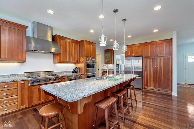 kitchen with dark wood finished floors, brown cabinetry, appliances with stainless steel finishes, wall chimney range hood, and a sink