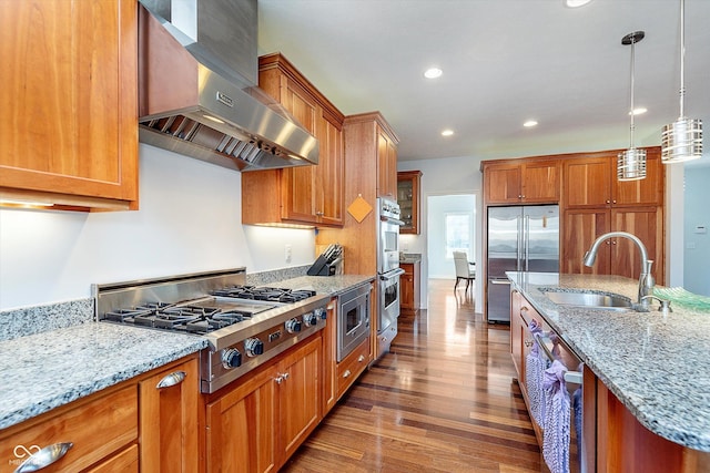 kitchen featuring brown cabinetry, dark wood-style flooring, built in appliances, wall chimney range hood, and a sink