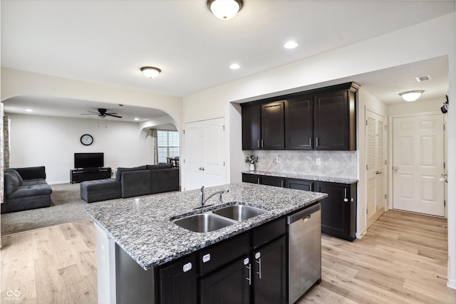 kitchen with light stone counters, arched walkways, a sink, an island with sink, and dishwasher