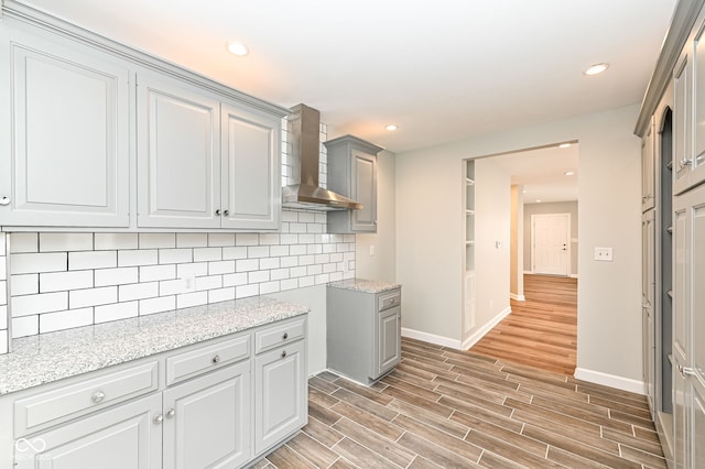 kitchen featuring light stone countertops, wood finish floors, gray cabinetry, wall chimney range hood, and tasteful backsplash