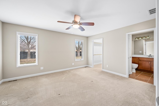 unfurnished bedroom featuring ensuite bathroom, light colored carpet, visible vents, baseboards, and a walk in closet