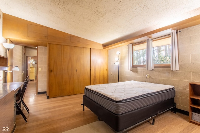 bedroom with concrete block wall, light wood finished floors, vaulted ceiling, a textured ceiling, and a closet