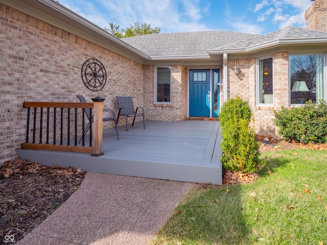 view of exterior entry with brick siding, a deck, and roof with shingles