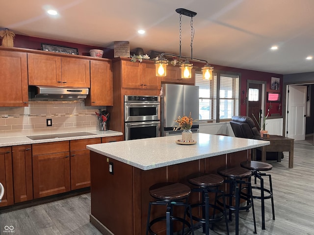 kitchen with under cabinet range hood, a kitchen bar, appliances with stainless steel finishes, and brown cabinetry