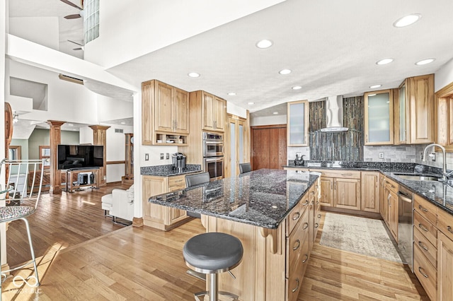 kitchen with light wood finished floors, appliances with stainless steel finishes, a sink, a kitchen island, and wall chimney exhaust hood