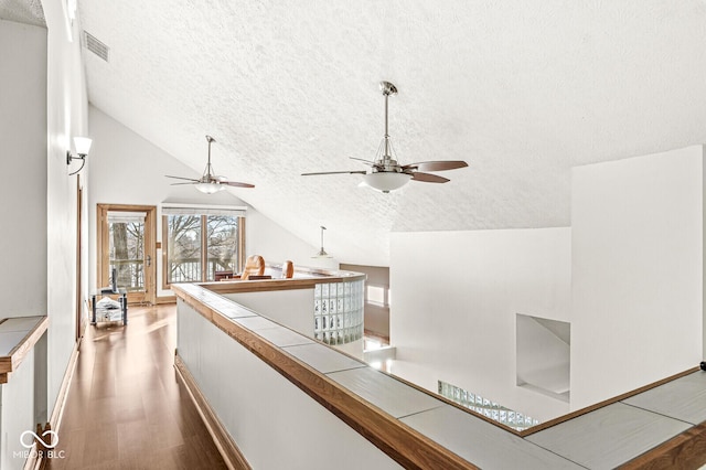 hallway with a textured ceiling, lofted ceiling, wood finished floors, visible vents, and an upstairs landing