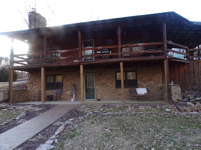 back of house with stone siding, a patio area, a chimney, and a balcony