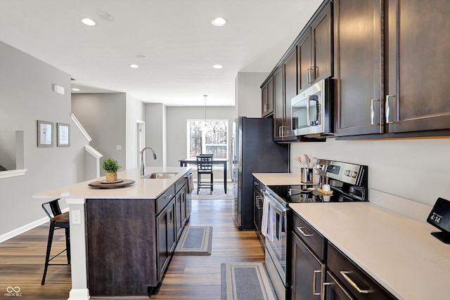 kitchen with an island with sink, a sink, stainless steel appliances, dark brown cabinetry, and dark wood-type flooring