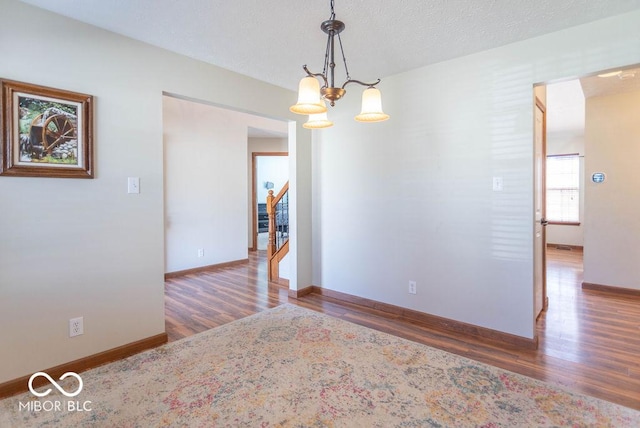 unfurnished room with a textured ceiling, wood finished floors, baseboards, stairway, and an inviting chandelier