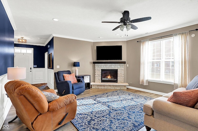 living room with crown molding, a ceiling fan, wainscoting, a stone fireplace, and wood finished floors