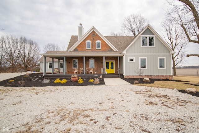 view of front facade featuring a shingled roof, a chimney, a porch, board and batten siding, and brick siding