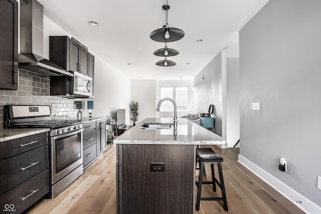 kitchen with a sink, wall chimney range hood, appliances with stainless steel finishes, backsplash, and an island with sink