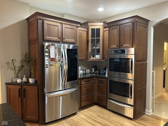 kitchen featuring arched walkways, stainless steel appliances, a warming drawer, dark stone countertops, and glass insert cabinets