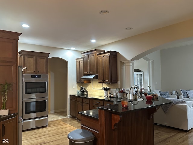 kitchen featuring arched walkways, a kitchen bar, double oven, under cabinet range hood, and ornate columns