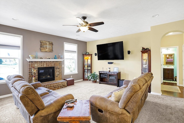 carpeted living room with a ceiling fan, arched walkways, a brick fireplace, and baseboards