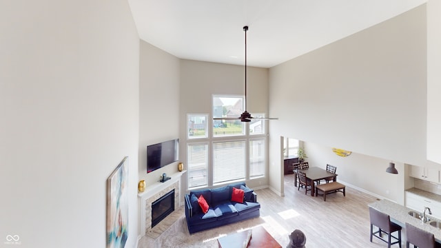 living area with light wood-style flooring, a high ceiling, a ceiling fan, a glass covered fireplace, and baseboards