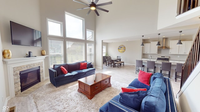 living area featuring ceiling fan, a stone fireplace, light wood-type flooring, baseboards, and stairs