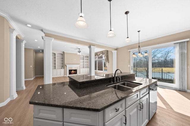 kitchen with a warm lit fireplace, ceiling fan, a sink, light wood-type flooring, and decorative columns