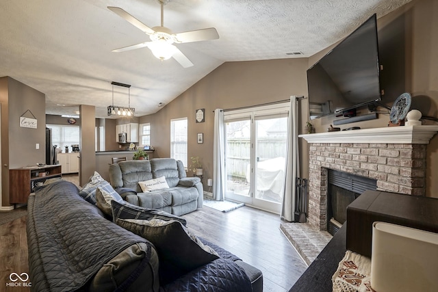 living area with a ceiling fan, wood finished floors, vaulted ceiling, a textured ceiling, and a brick fireplace