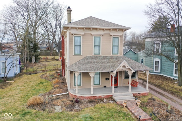 italianate home featuring roof with shingles, a chimney, covered porch, fence, and a front lawn