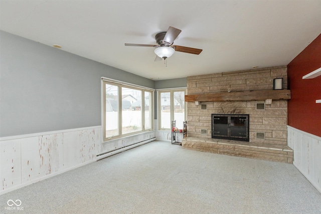 unfurnished living room with a wainscoted wall, a baseboard heating unit, carpet flooring, ceiling fan, and a stone fireplace