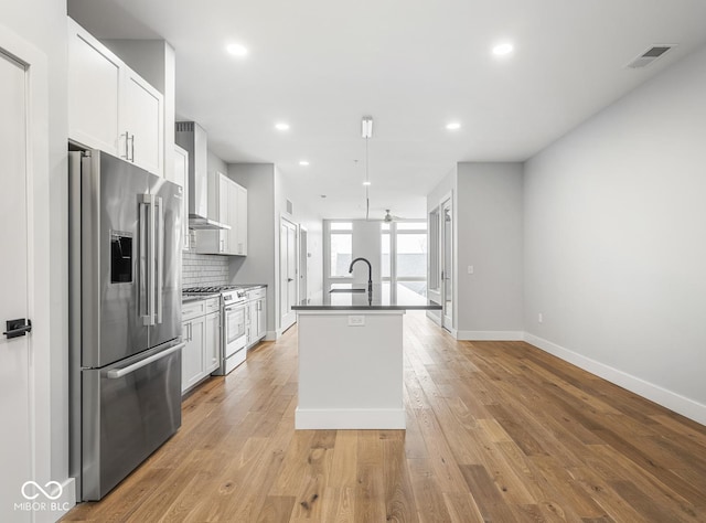 kitchen featuring a center island with sink, dark countertops, wall chimney exhaust hood, appliances with stainless steel finishes, and backsplash